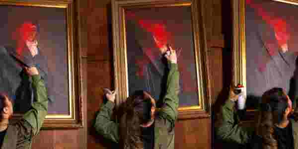 We see a woman from behind spraying an oil painting portrait of probably British aristocracy with red spray can , slashes throguh it, frames in gold infront of wooden wall