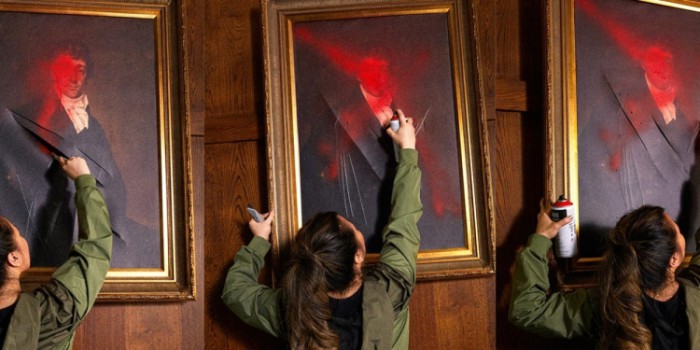 We see a woman from behind spraying an oil painting portrait of probably British aristocracy with red spray can , slashes throguh it, frames in gold infront of wooden wall