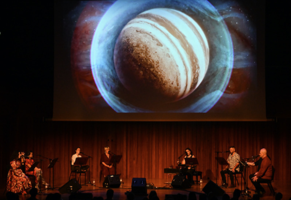 We see a large image of a planet on a screen in the background of the stage at Milton Court and the eight singers from Roomful Of Teeth sitting in front of it on chairs with indivisual mic stands and mics.